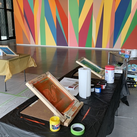 Various screenprinting supplies are set up in the Stanley Museum of Art lobby. These supplies have been used--ink jars are open, and the screens are flipped up. One screen has an orange ink coating it, and another screen has a greenish blue ink.