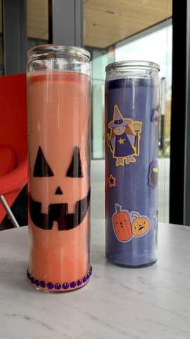 An example of the candle craft--one orange candle with black marker draw on it so it looks like a jack-o-lantern, and one purple candle decorated with rhinestones and Halloween stickers of a witch and a pumpkin.