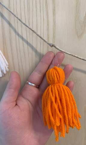 A person holds their hand up to add emphasis to part of a garland; an orange ghost shaped yarn pompom or tassel is attached to a length of twine with clear fishing line.