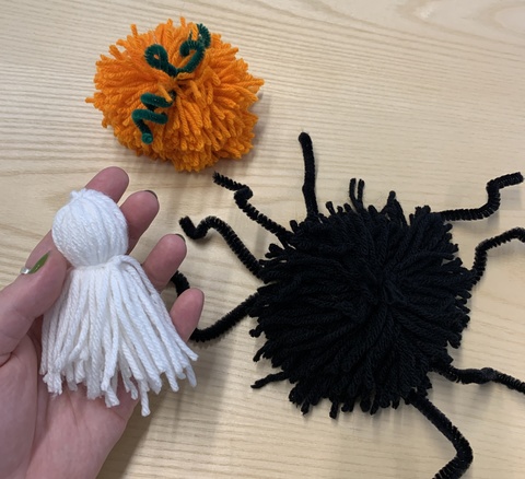 A photo displaying three times of Halloween yarn pompoms: a white ghost, an orange pumpkin, and a black spider.