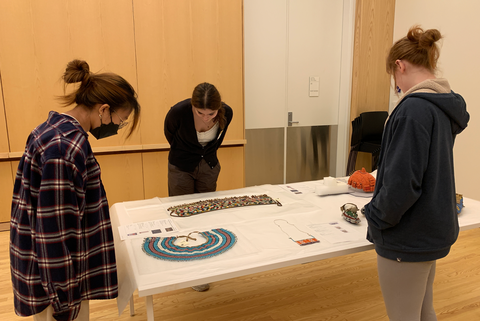 Three students stand in the Visual Classroom, looking down at a display of artwork from the collection that is laid out for them on the table.