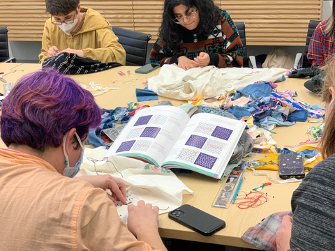 Students are gathered around a conference table, each working on their own mending project. One student in the foreground, back to the camera, is focused on a book that is open, displaying various sashiko stitching techniques and patterns.