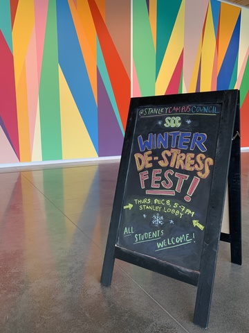 A photograph of a chalk sandwich-board style sign. It is placed in front of the Stanley's lobby mural. In colorful letters, the sign reads: "SCC Winter De-Stress Fest! Thursday, December 8, 5 - 7 pm. All students welcome!"
