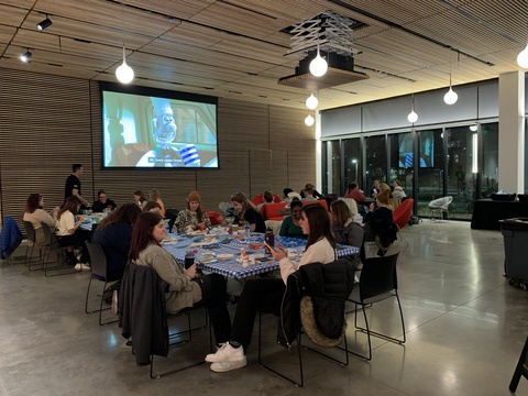 A photography of the lobby, filled with students there for the event. The lobby is dimly lit, and the projector at the far end is showing Wallace and Grommit. There are clusters of tables with students grouped around them, eating, crafting, and hanging out.