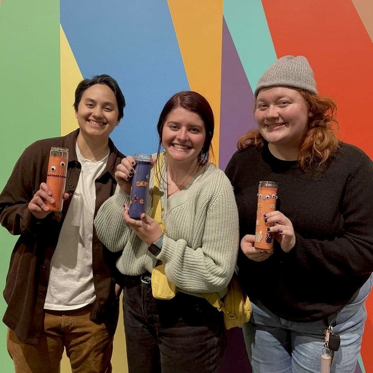 Three students pose in front of the Odita mural in the Stanley lobby with their crafts: votive candles decorated with stickers, rhinestones, and markers.
