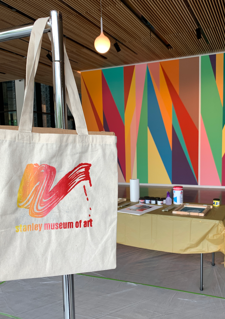 A photo of one of the tote bags, hanging off a metal coat rack. In the background are screenprinting supplies set up in the Stanley lobby with the Odita mural in the background.