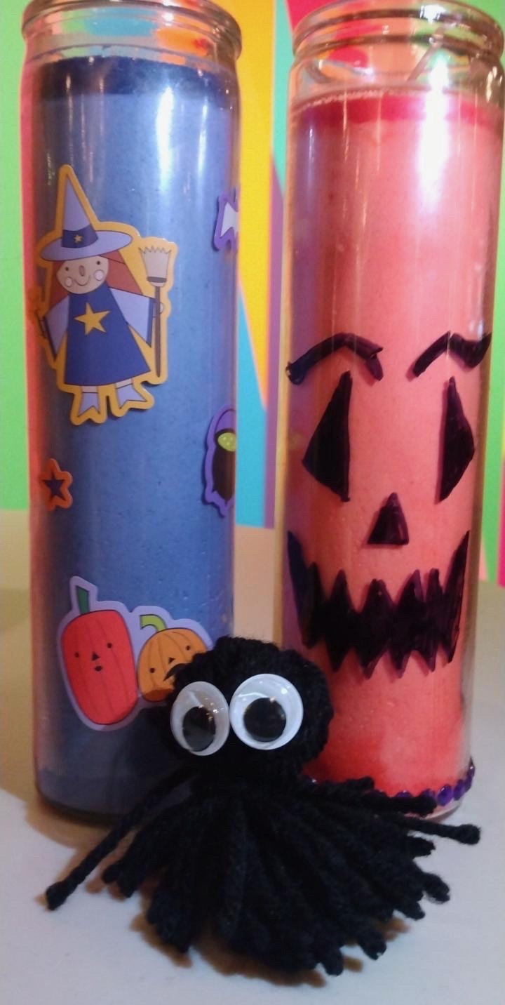 An example of some of the crafts made by student attendees. Two tall votive candles, one orange with a jack-o-lantern face drawn on it, and one purple and covered in stickers, sit on a table with a black pompom ghost with googly-eyes in front of it.
