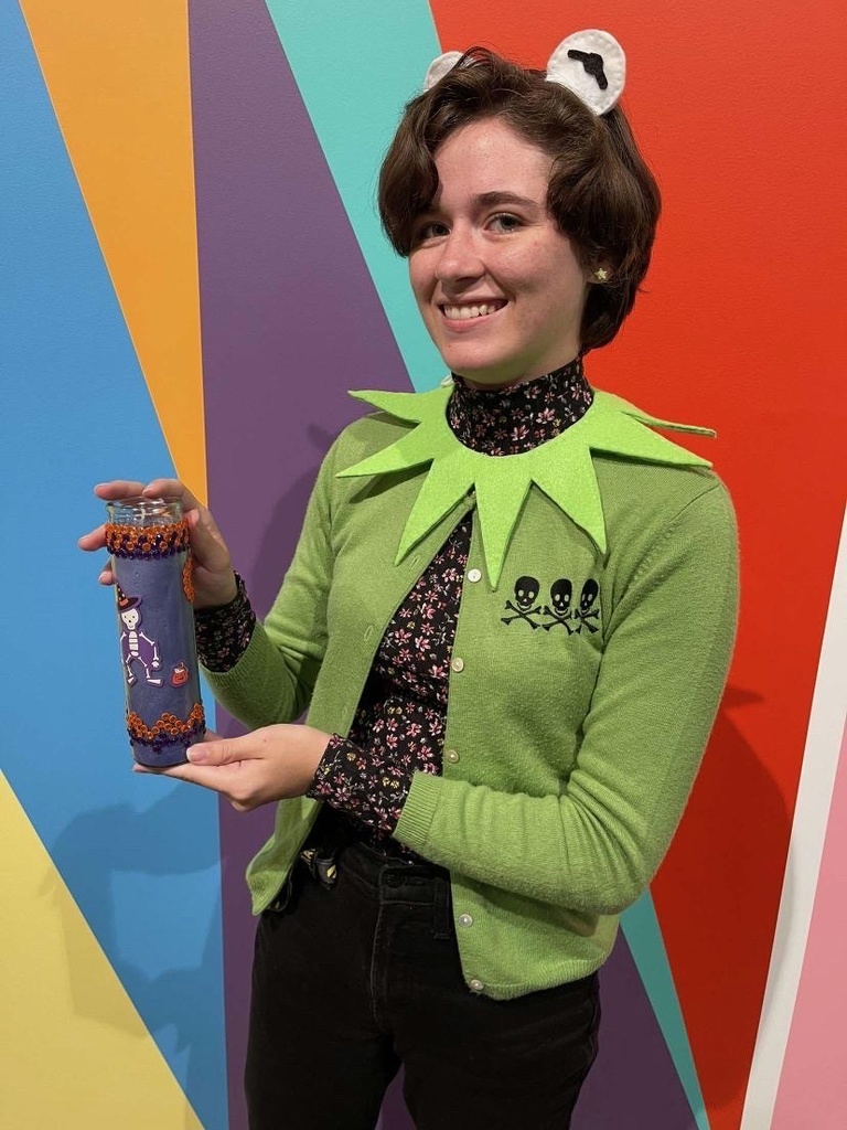 A student, in a homemade Kermit the frog costume, poses in front of the Odita mural in the Stanley lobby with their craft: a tall purple votive candle, decorated with stickers and rhinestones.
