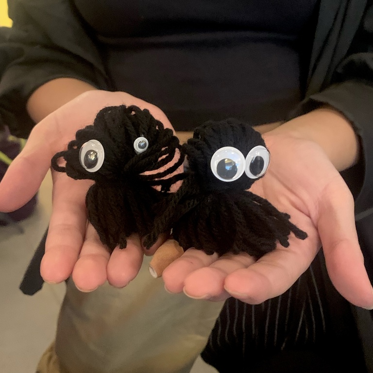 A student holds their crafts in their hands: two black yarn pom-pom creatures with mismatched googly eyes.