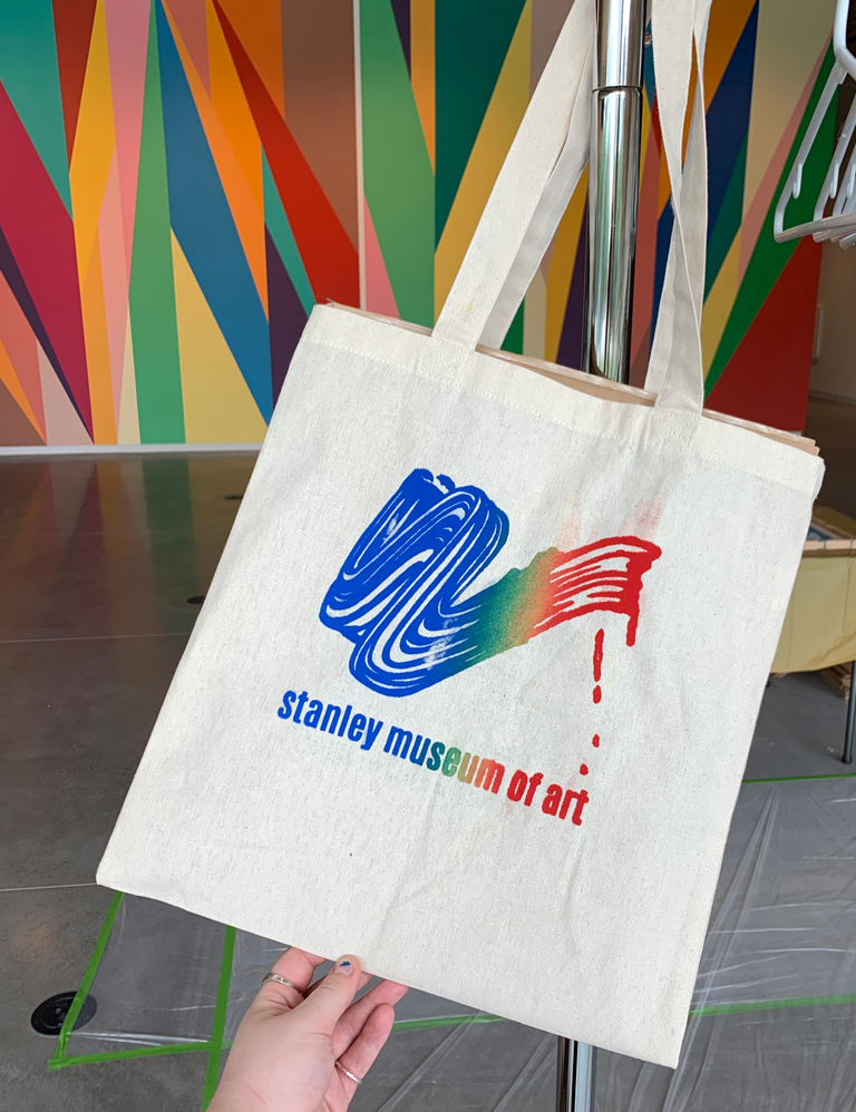 A photo of a hand holding a freshly printed tote bag that has been printed in a rainbow gradient of colors. In the background is the Odita mural in the Stanley lobby.