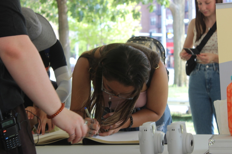 A guest bends over a table in Gibson Square Park that holds the guest book, and signs her name next to a Polaroid picture taken of her.