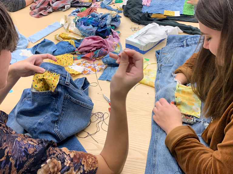 An image of two students sitting together, both bent over their work. The one on the left pulls thread through a garment that they are patching. The one on the right adjusts and makes stitches into the garment she is mending; a pair of ripped jeans with a yellow floral patch she's added.