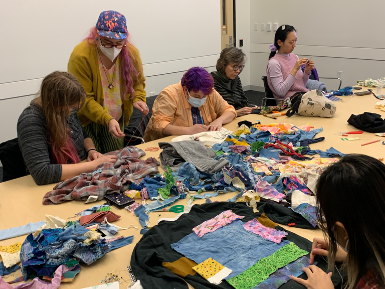 A shot of the table in the seminar room. it is covered in colorful bits of scrap fabric for people to use to patch their garments. Five people sit across the table, facing the camera. One person is in front of the camera, facing away, looking at the garment that they are mending.