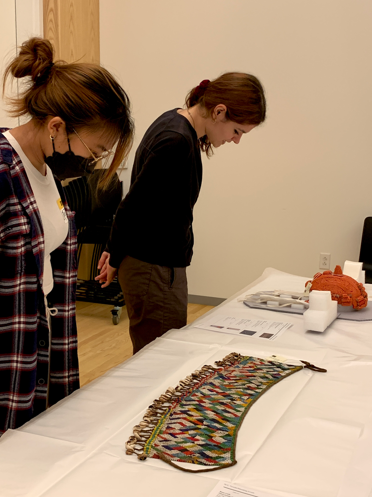 Two students gaze down at work from the collection that is displayed on the white tables in front of them. One in the background looks at a red beaded Yoruba lawyer's wig. The one in the foreground gazes at a beaded apron.