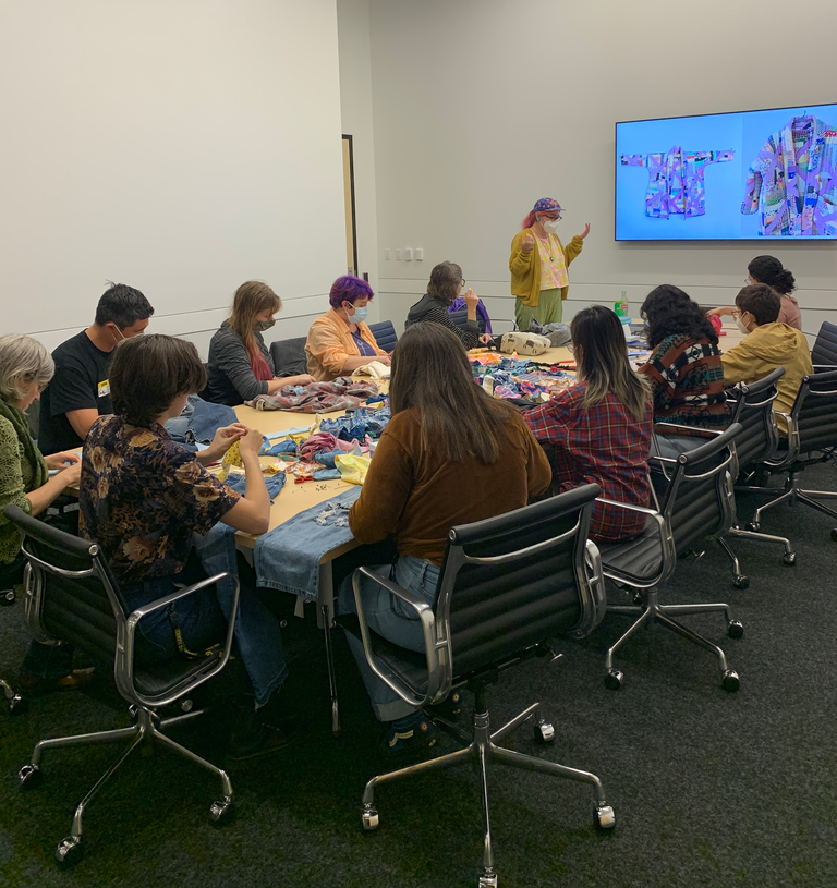 An image of Lilah leading the sashiko workshop. She stands at the head of the table, gesturing towards a screen which displays images of her work that makes use of sashiko. 10 or 15 students sit around the table, which is covered in fabric scraps and their mending projects, watching her.