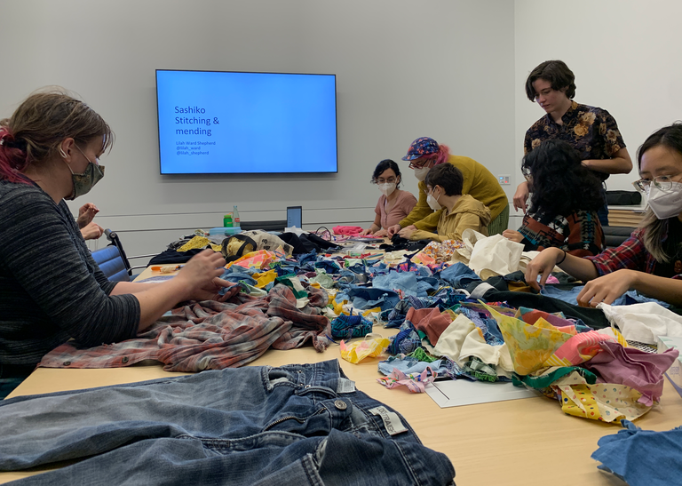 Students begin working on their mending projects. Everything is laid out on the table--scraps of fabric, sewing needles, ripped garments, and threads. 