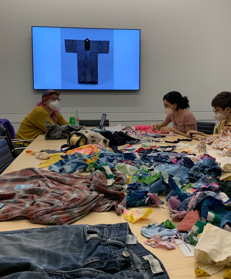 Students begin working on their mending projects. Everything is laid out on the table--scraps of fabric, sewing needles, ripped garments, and threads. The screen in the background at the head of the table displays an example of a coat decorated with sashiko statching.