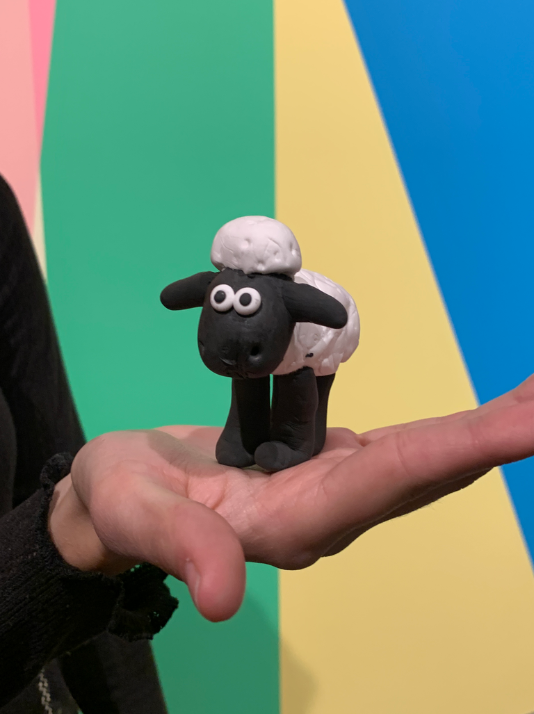 Another clay creation: a student holds in their hand a replica of Shaun the Sheep, from Wallace and Grommit, with the lobby mural in the background. 