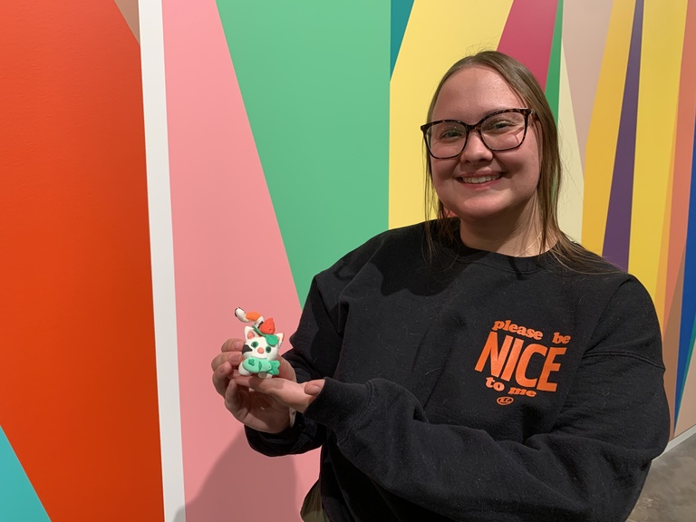 A student poses, smiling, in front of the lobby mural with their clay creation: a calico colored cat with green eyes, a strawberry hat, and a fish in its mouth. 