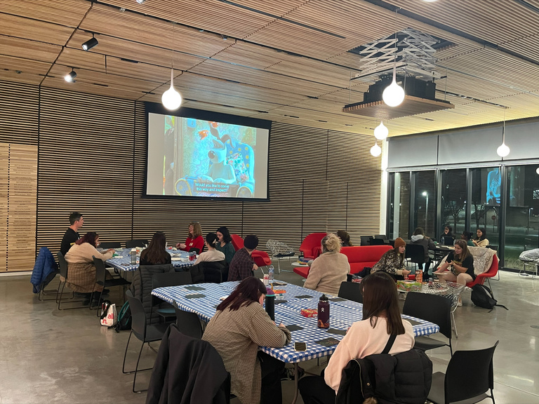 A shot of the lobby, full of students crafting, eating, and hanging out. A large screen at the far end of the room is playing Wallace and Grommit.