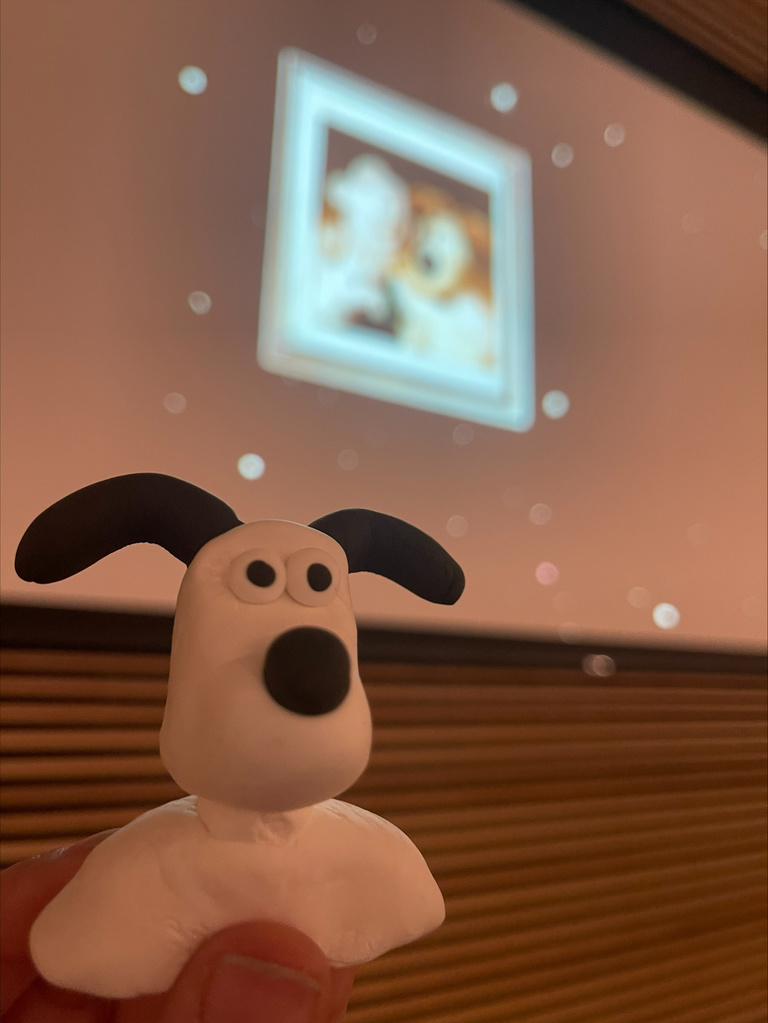 A hand holds up a clay replica of Grommit the dog. In the background is the projector screen, which is playing the credits for Wallace and Grommit.