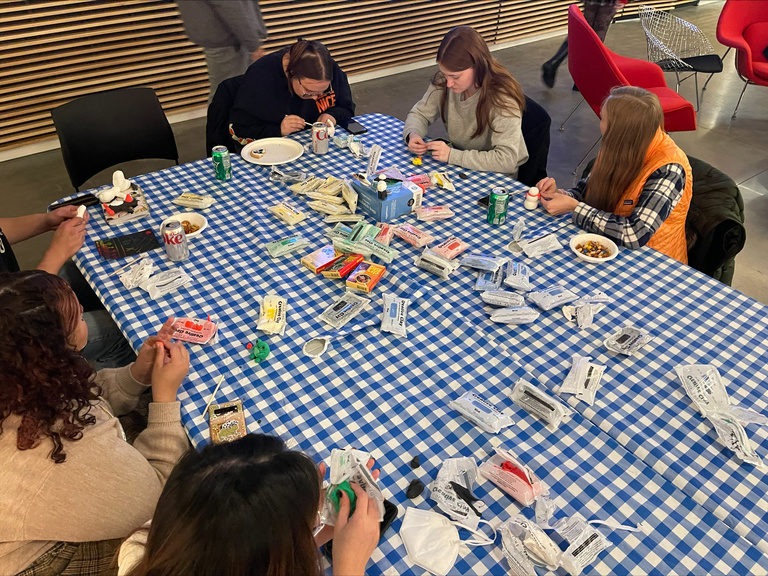 A photo taken from above, looking down at one of the craft table. Students sit around a table with a blue gingham tablecloth, sculpting various things out of air dry clay.
