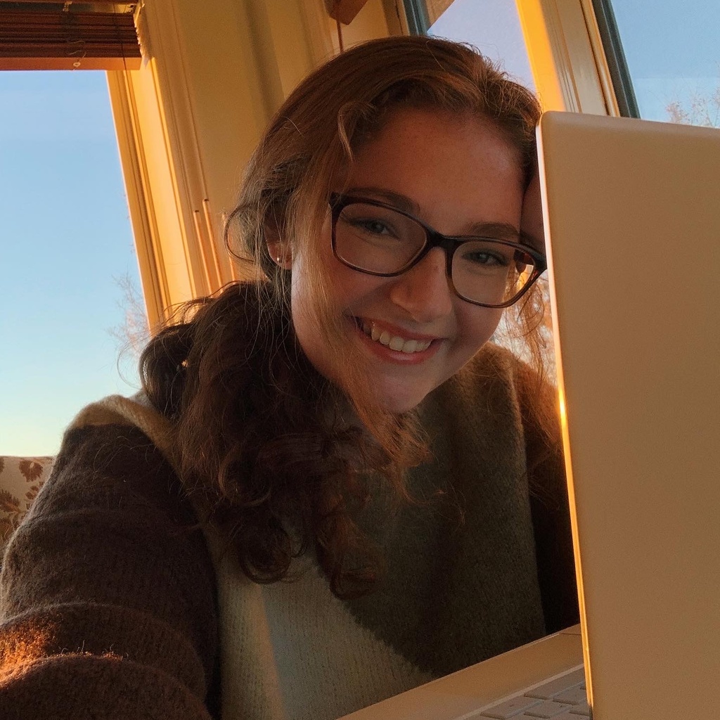 A selfie of Emalie; she is sitting with her laptop during golden hour. She stares at the camera, smiling cheerfully. She has curly, reddish hair and dark glasses.