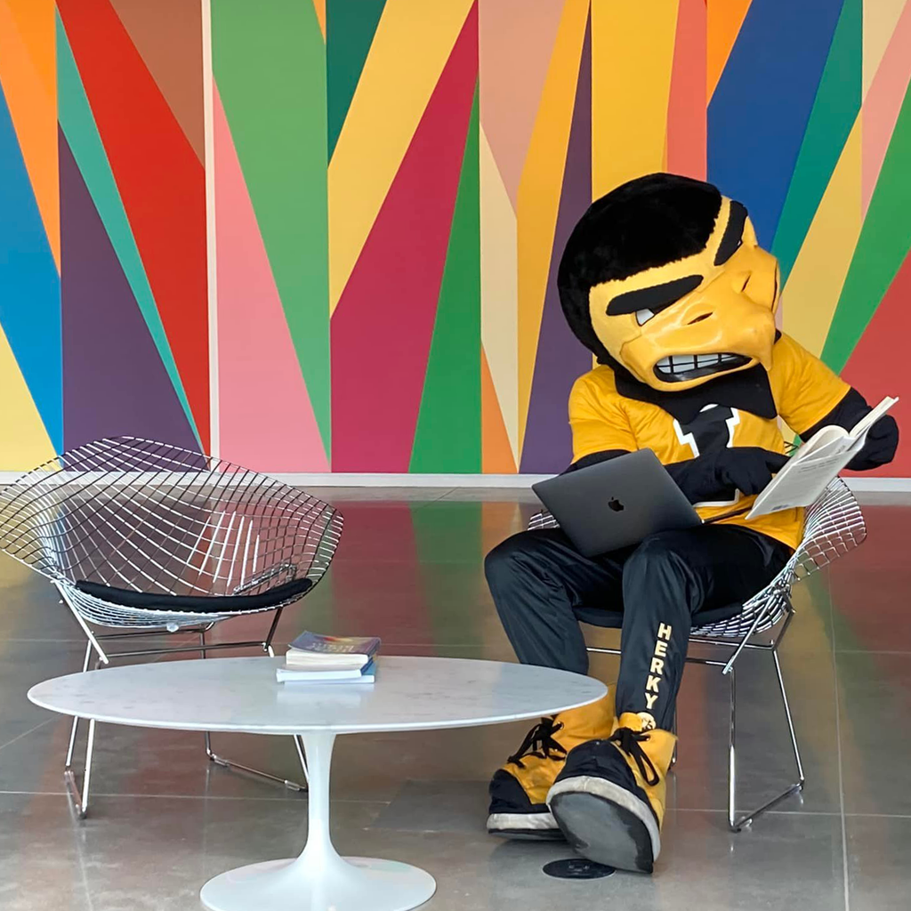 Herky the mascot sits at one of the tables in the Stanley Museum of Art lobby. He is studying, balancing a laptop on his lap and flipping through a book in his hands.