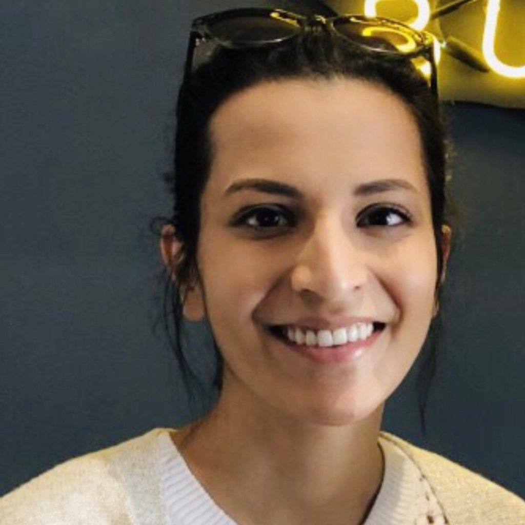 Akansha smiles at the camera; her dark hair is pulled back behind her head, and she has sunglasses resting atop her head. She is wearing a white shirt and is placed against a dark gray background.