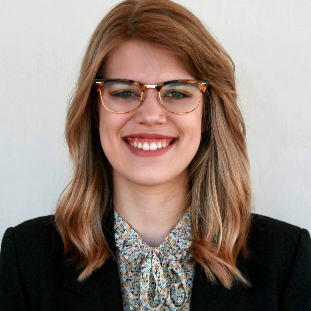 A smiling student; dressed in a black blazer with a floral collared shirt, with shoulder length red-blonde hair, and tortoiseshell glasses.