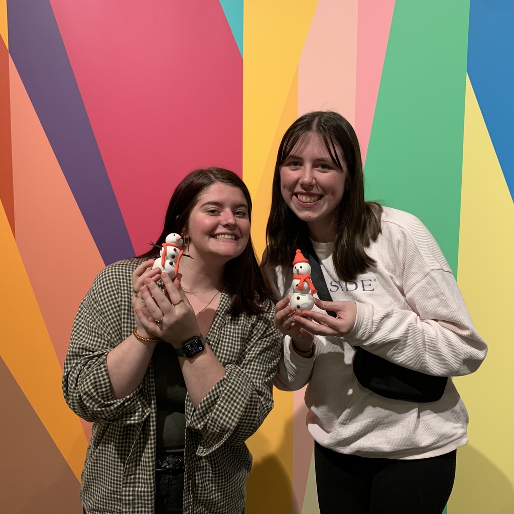 Two students stand together in front of the Odita mural in the Stanley lobby. Each of them is holding their clay creation in their hands. They're smiling, posing with the two clay snowmen they've made.