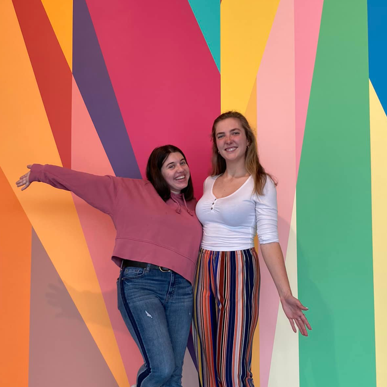 Two students embrace, posing in front of Odili Donald Odita's mural entitled "Surrounding" in the Stanley Museum of Art lobby.
