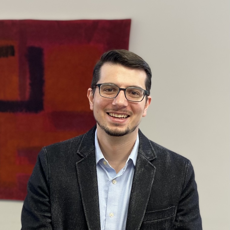 A professional headshot photo of a smiling man with dark hair, standing in front of a white wall with a red painting. 