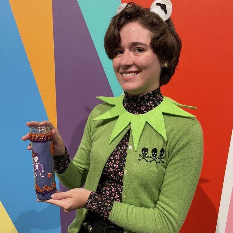 A student, in a homemade Kermit the frog costume, poses in front of the Odita mural in the Stanley lobby with their craft: a tall purple votive candle, decorated with stickers and rhinestones.