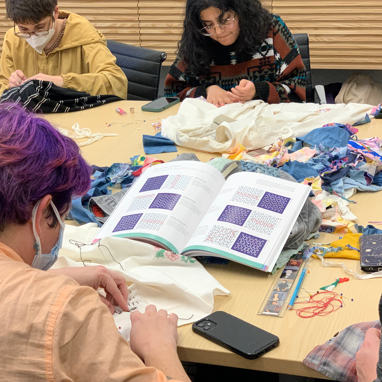 Students are gathered around a conference table, each working on their own mending project. One student in the foreground, back to the camera, is focused on a book that is open, displaying various sashiko stitching techniques and patterns.