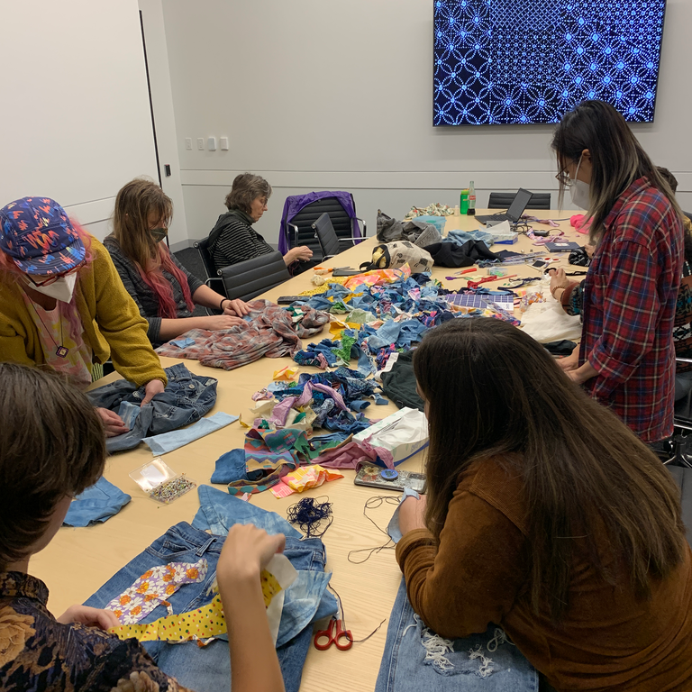 A photo of the class gathered at the table. Fifteen or twenty students sit at a long conference table covered in fabric scraps, books, and garments. At the head of the table is a screen that is displaying a close up of varying sashiko stitches.