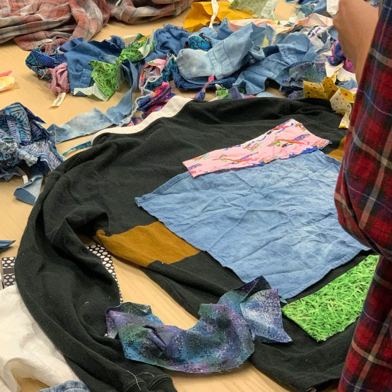 A close up example of a garment one student works on--it is a black crewneck sweater that she has covered with patches of varying fabrics, achieving a patchwork look.