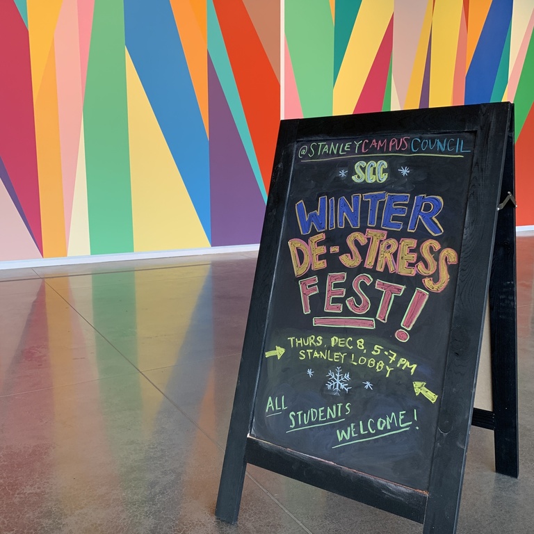 A photograph of a chalk sandwich-board style sign. It is placed in front of the Stanley's lobby mural. In colorful letters, the sign reads: "SCC Winter De-Stress Fest! Thursday, December 8, 5 - 7 pm. All students welcome!"