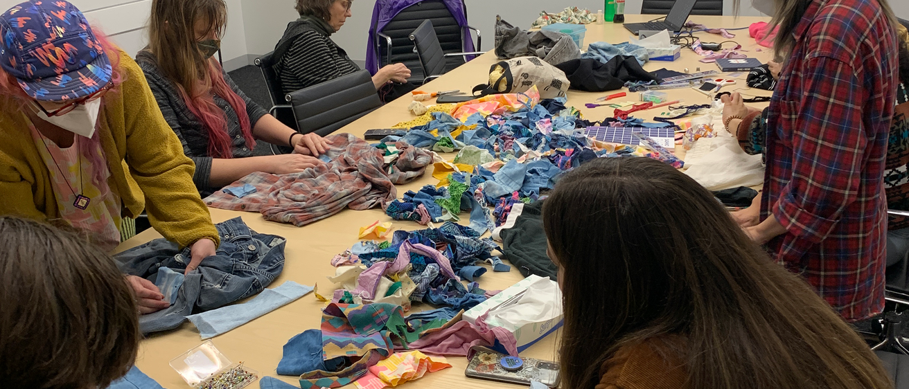 A photo of the class gathered at the table. Fifteen or twenty students sit at a long conference table covered in fabric scraps, books, and garments. At the head of the table is a screen that is displaying a close up of varying sashiko stitches.