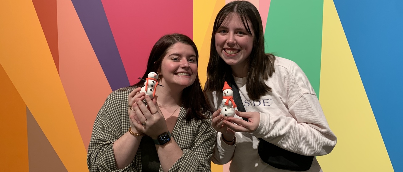 Two students stand together in front of the Odita mural in the Stanley lobby. Each of them is holding their clay creation in their hands. They're smiling, posing with the two clay snowmen they've made.